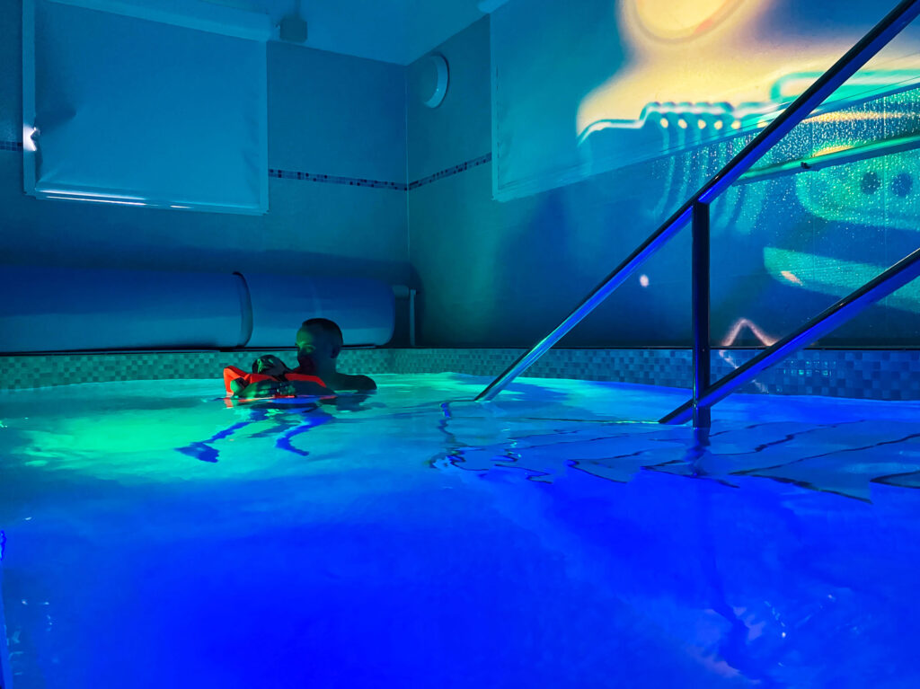 A father and his son, who is wearing a floating vest, are in the hydrotherapy pool at Russell House. There is calming blue low lighting with patterns being projected onto the wall.
