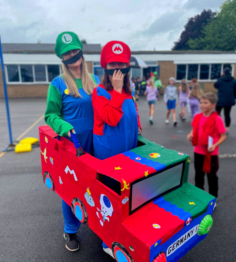 Two female primary school teachers dressed up as Mario and Luigi for a fundraising event. They are both stood inside a cardboard car they are holding up to their waists which has been decorated with Mario designs.