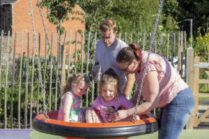 A mum and dad are helping their two daughters to settle onto a large round swing in the garden at Russell House.