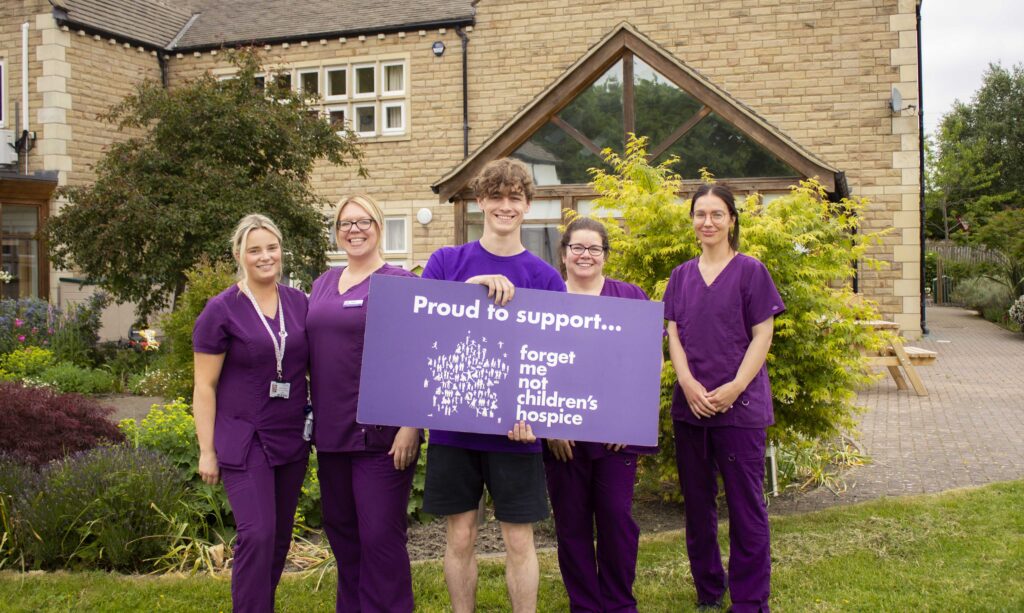 Rhys Connah stood in the middle of 4 female care staff in the garden of Russell House. He is holding a large sign saying "proud to support Forget Me Not Children's Hospice"