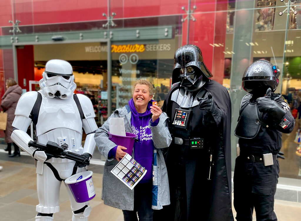 Volunteer Linda holding a collection bucket as she is stood between a Stormtrooper and Darth Vader at a fundraising event at Trinity Walk