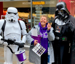 Volunteer Linda holding a collection bucket as she is stood between a Stormtrooper and Darth Vader at a fundraising event at Trinity Walk