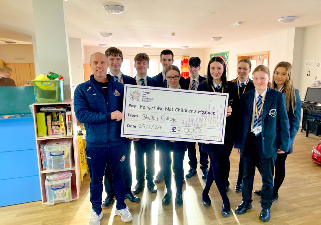 A group of high school children from Shelley College wearing uniforms and a male teacher stood in the Hub. They are holding a large cheque donation of £2000.