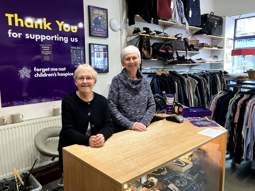 Two older female volunteers stood behind the counter in the Holmfirth shop. The sign behind them read "thank you for supporting us"
