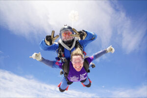 A forget me not fundraiser in the sky, doing a tandem skydive with an instructor on their back. They are both smiling at the camera.
