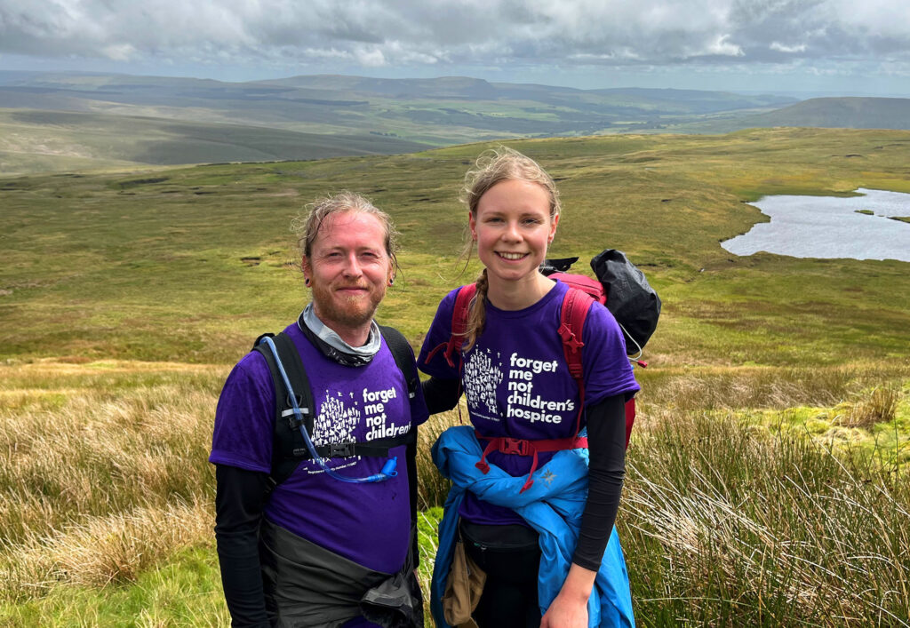 A man and woman, both wearing a purple forget me not t-shirts and backpacks, stood in the Yorkshire countryside while doing the Three Peak challenge.