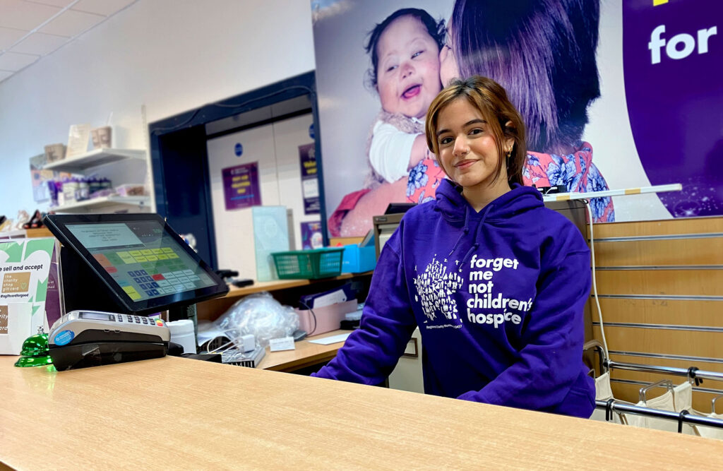 A young Asian retail employee wearing a purple Forget Me Not hoodie stands behind the counter at the Manchester Road store.