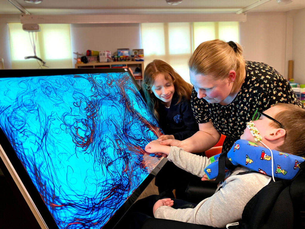 A young white boy in a wheelchair is sat in front of a large interactive touchscreen which has patterns on it. His mum is beside him holding his hand to the screen to help him, while his sister looks on smiling.