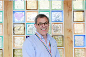 Jeremy, A Forget Me Not Trustee, stood in front of the brightly coloured wall of glass blocks of the memory wall at Russell House