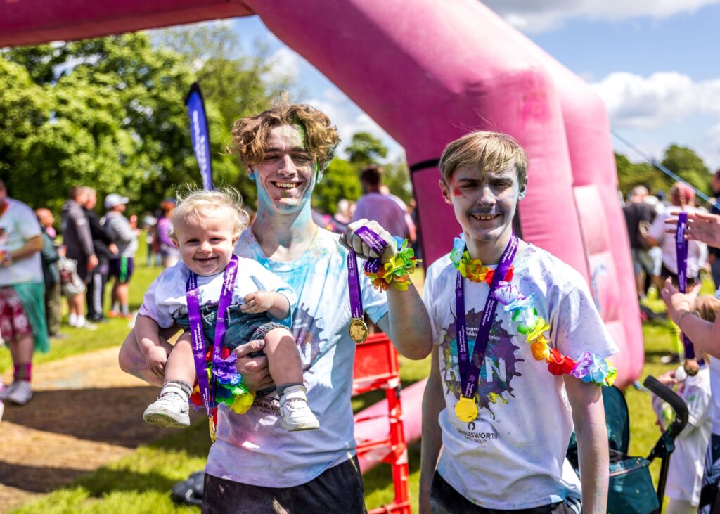 Ambassador Rhys Connah at the Forget Me Not colour run with his brothers. He is holding his baby brother in his arm as they all wear medals and are covered in colourful paint powder.