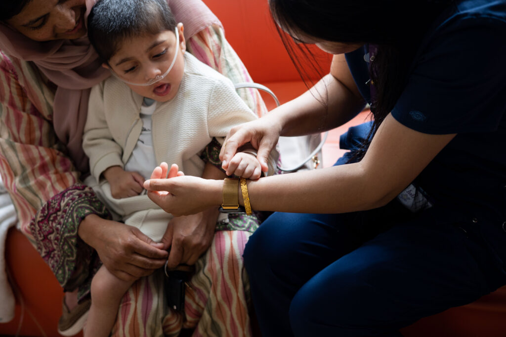An Asian woman with her son on her knee sat next to an Asian member of the family support team, who is holding the sons hand to her wristwatch. The son has a breathing tube in his nose.