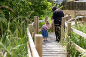 A white woman wearing a black dress with brown hair holding her young daughters hand as they stand on a boardwalk through a grassy garden area