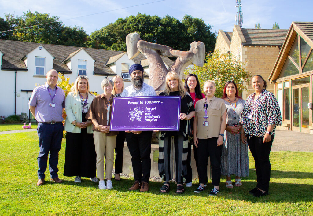 Representatives from Kirklees College with staff members from Forget ME Not stood holding a "proud to support Forget Me Not" sign in front of the statue in Russell House gardens