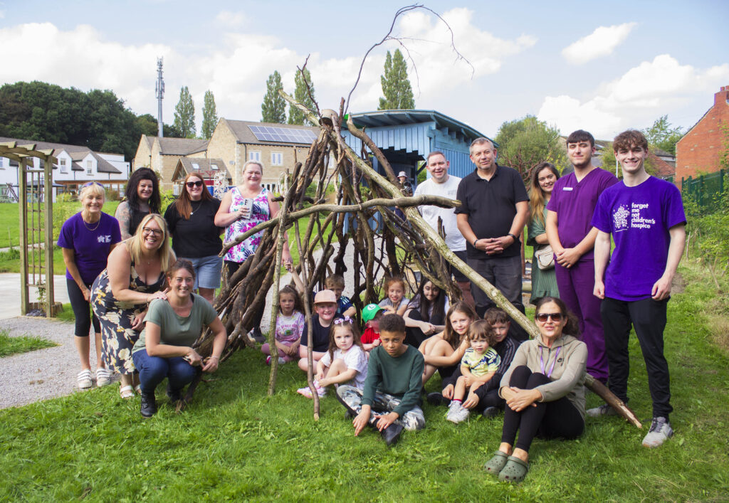 A group of Forget Me Not staff, families and ambassador Rees Connah in the garden at Forget Me Not. They are gathered round a shelter made out of twigs and branches which was created at the bushcraft activity day.