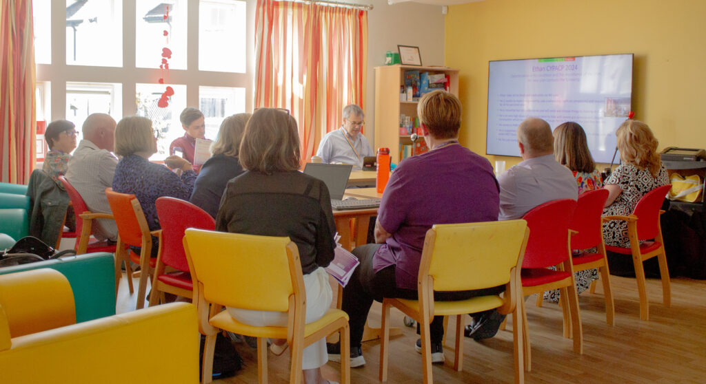 Members of the Forget Me Not leadership team sat around a table in the Hub. There is a large screen showing a presentation which they are all watching.