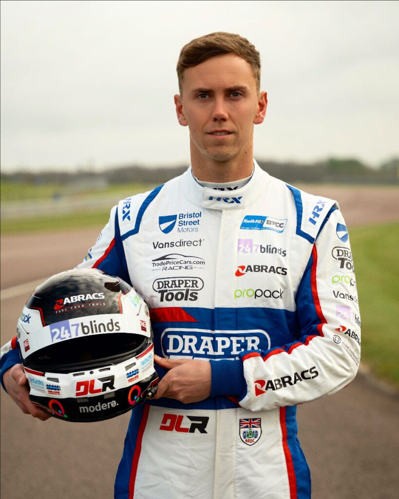 Racer Daniel Lloyd holding his helmet stood on a race track wearing his racing suit