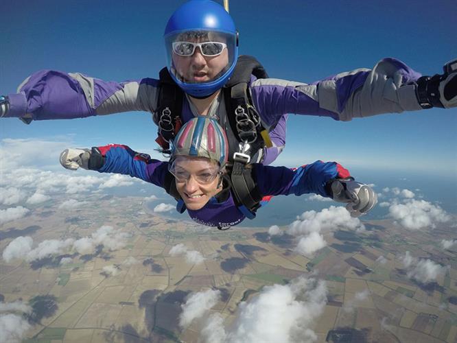 Two skydivers, male and female, smile at camera while freefalling with the ground below them.