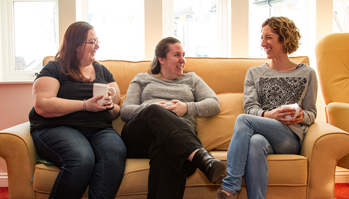 Three women sat on a sofa in disscusion drinking coffee.